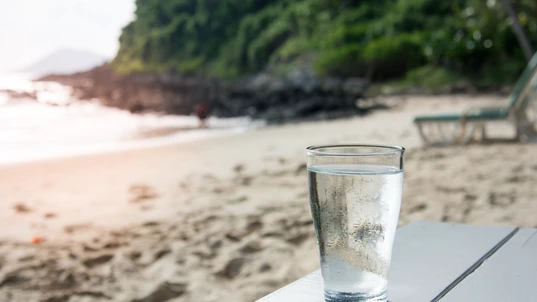 A glass of cold water on white wooden table on the beach. Stay hydrate this summer.