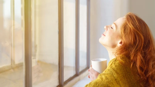 Blissful young woman enjoying the sun on her face as she leans on a window sill with her head tilted back and a mug of coffee