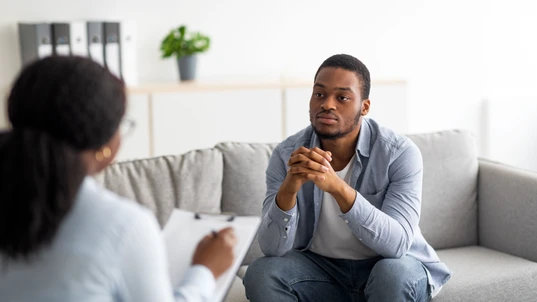 Psychological help service. Depressed black male patient having psychotherapy session with counselor at mental health clinic. Young man with emotional problems consulting professional therapist