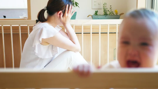 Woman with head in her hands next to cot with crying baby inside